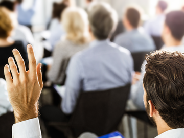 Die nächsten Veranstaltungen Mann hebt linke Hand zu einer Wortmeldung während einer Infoveranstaltung