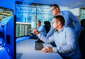 Security-Operation-Center Zwei Männer schauen am Arbeitsplatz auf Monitore, ein dritter sitzt im Hintergrund und telefoniert