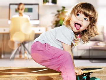 Homeoffice Kind spielt mit einem Skateboard, im Hintergrund die Mutter wie sie im Homeoffice arbeitet